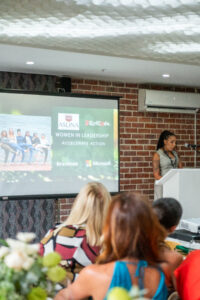 woman in a grey outfit speaking at the Accelerate Action: A Creative Brunch for Women in Business Leadership function. On the slideshow behind her we see the heading "women in leadership. accelerate action".