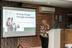 woman in a multicoloured outfit speaking at the Accelerate Action: A Creative Brunch for Women in Business Leadership function. On the slideshow we see the heading "Driving Change Through Innovation"