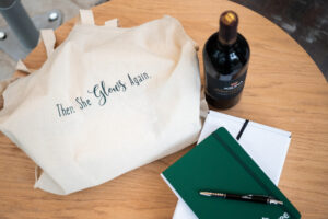 table topped with a wine bottle, a notebook and pen, a white box and a tote bag with the words "there she glows again."