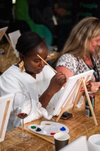 Women fat the Accelerate Action: A Creative Brunch for Women in Business Leadership function, painting on small canvasses and having wine - 3