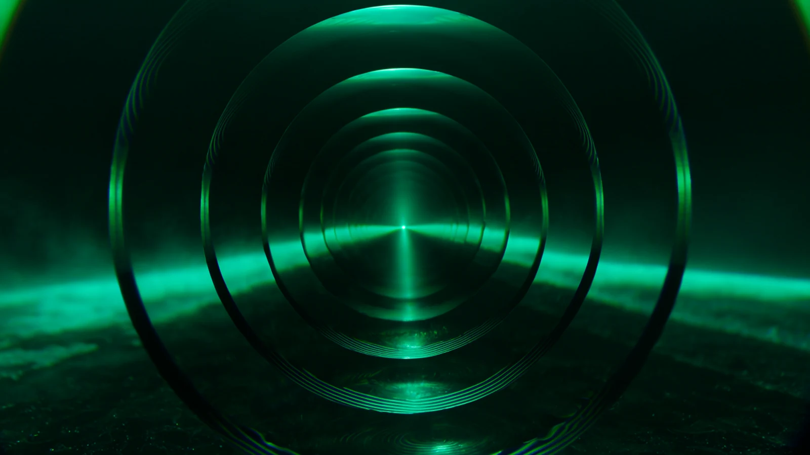 Concentric precision lenses stacked one behind another in dark space, green light refracting through each layer into rings.